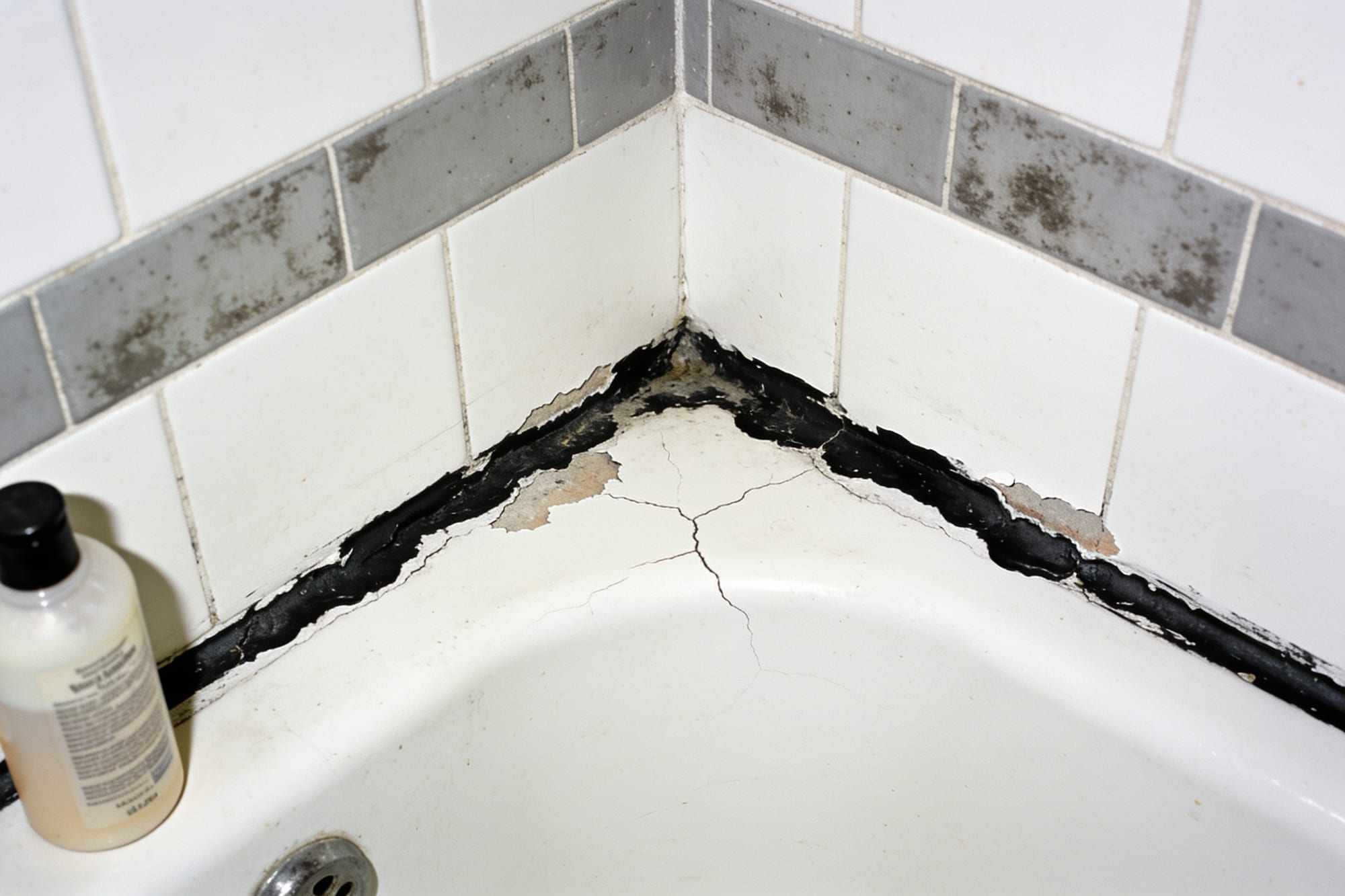 A bath edge with cracked, black, mouldy sealant and grey grout
