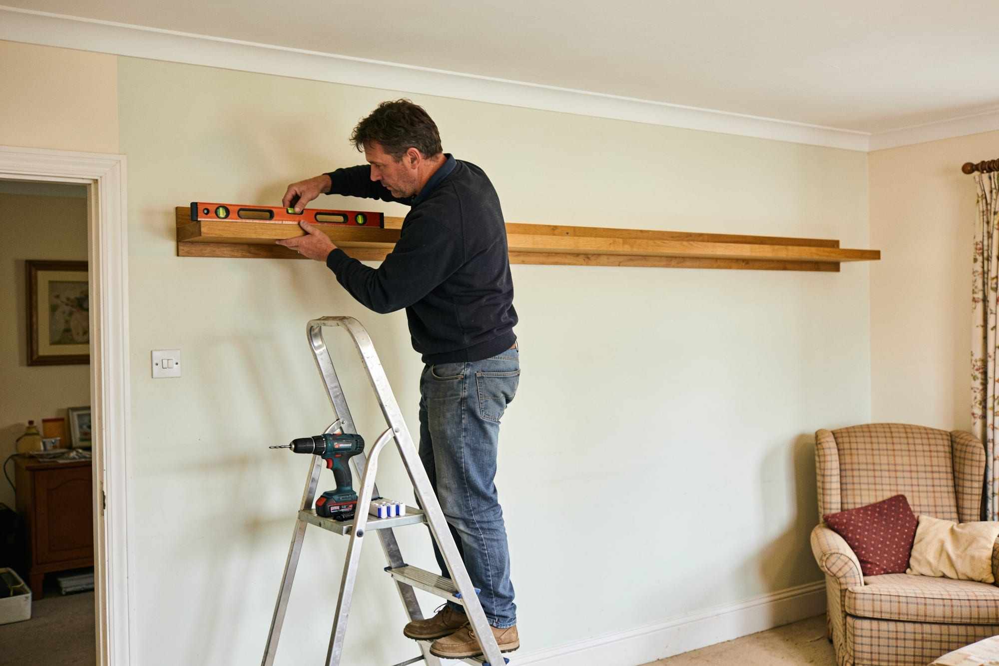 A handyman on a step ladder levelling a long oak shelf on a living room wall